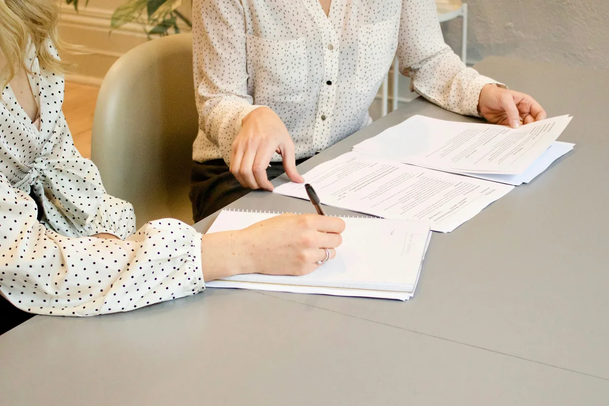 Two people at a meeting table, one writing notes while the other points to printed documents