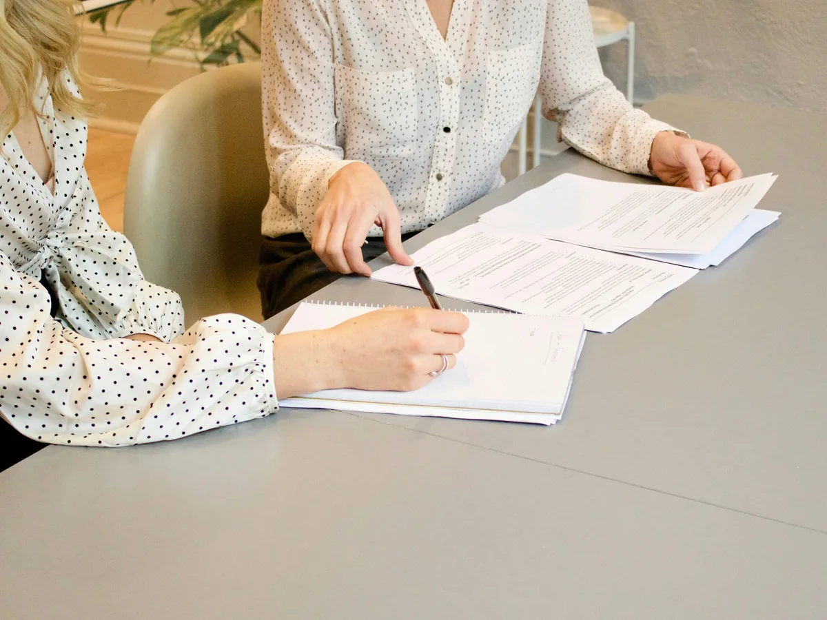 Two people at a meeting table, one writing notes while the other points to printed documents