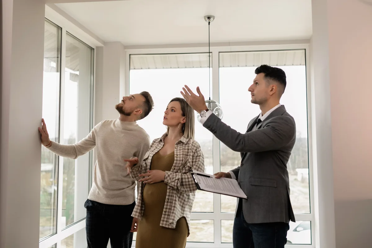 A real estate agent with a clipboard shows a young expecting couple through a bright, empty home