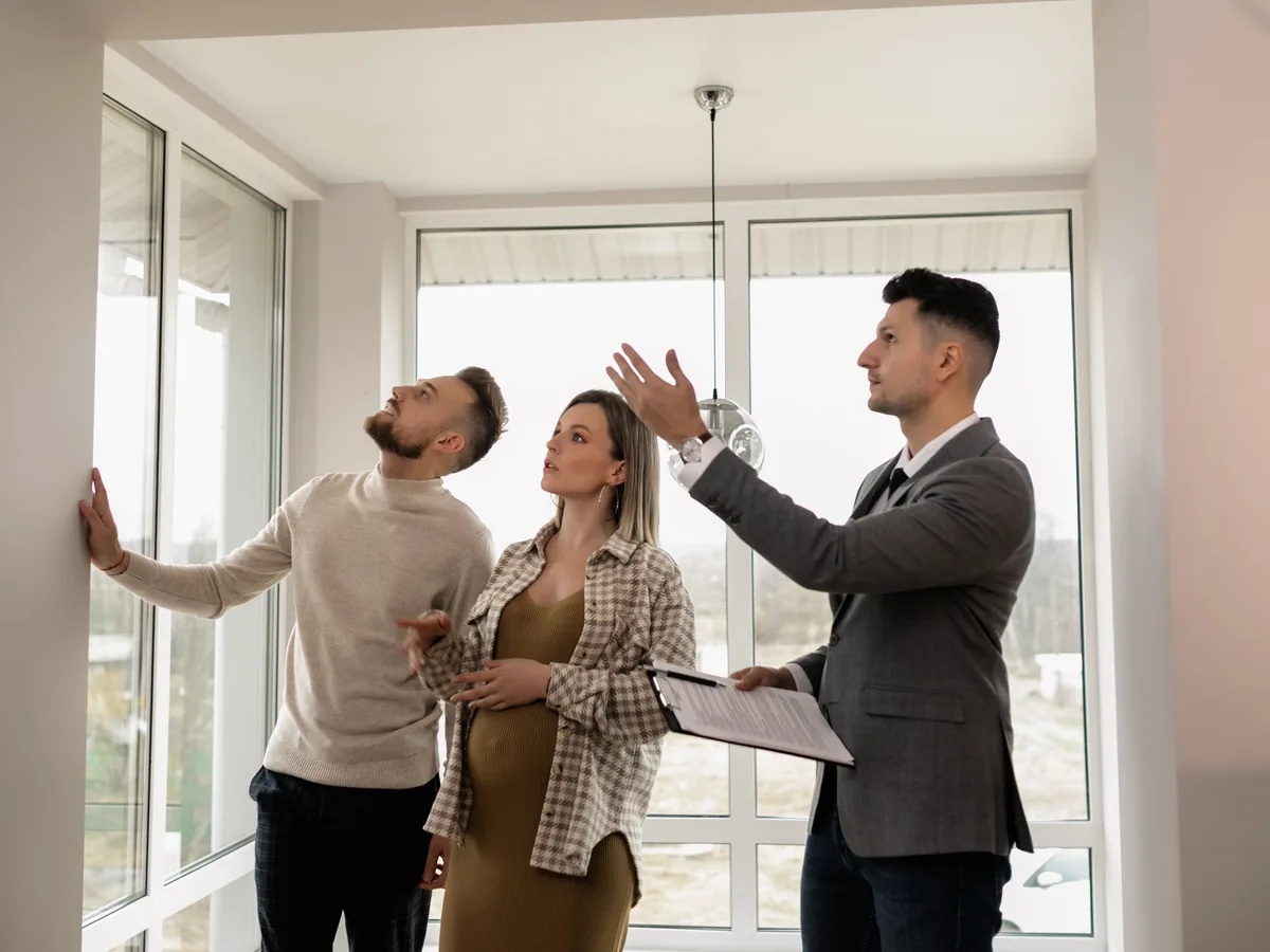 A real estate agent with a clipboard shows a young expecting couple through a bright, empty home