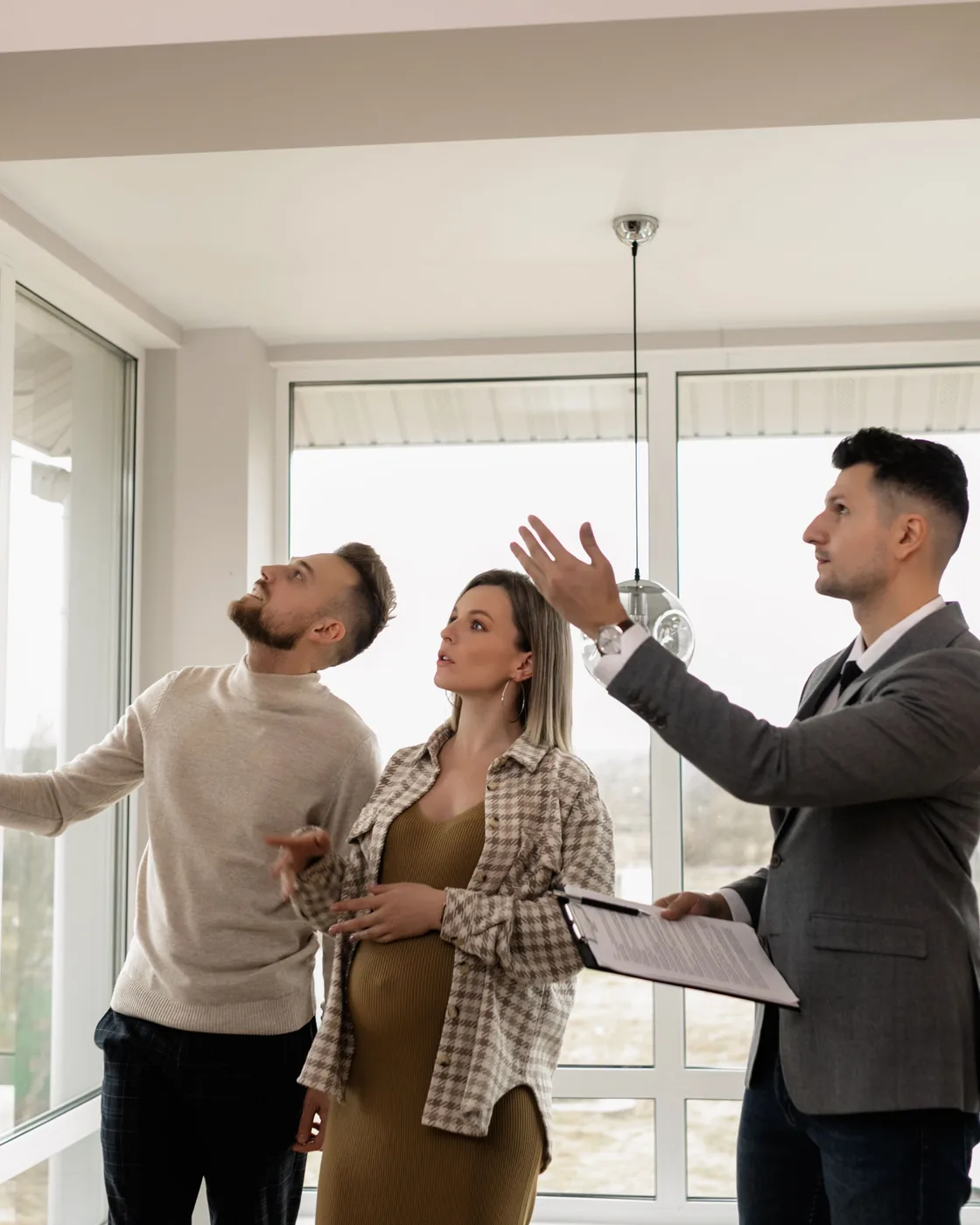 A real estate agent with a clipboard shows a young expecting couple through a bright, empty home